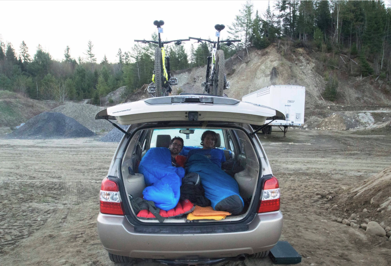 Two riders in sleeping bags in the back of a car with mountain bikes on the roof